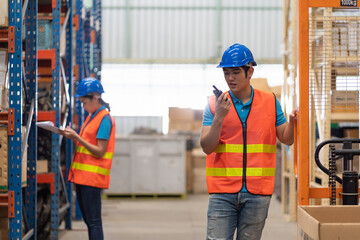 Asian young male employee factory worker with safety vest and helmet working and using communication radio in automotive spare parts warehouse