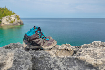 Fototapeta na wymiar Close up of pair of old worn out weathered hiking boots on a rocky cliff over turquoise water of Georgian Bay, sunny day, selective focus, space for copy. Hiking, camping, active lifestyle concept.