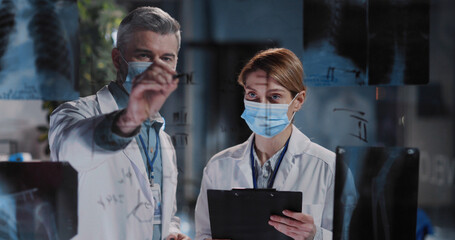 Flu Epidemic. Caucasian Adult Doctor with Female Assistant Executive in Face Masks Discussing Lung Disease and X-ray Photographs in Medical Office Laboratory Hospital. - obrazy, fototapety, plakaty