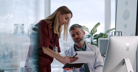 Obraz premium Young Caucasian Nurse Woman Asking Advice from Adult Doctor Negotiating in Private Medical Office. Staff Cooperation. Mixed Ethnicity. Hospitals.