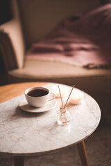 Liquid home fragrance with glass bottle and wooden sticks, cup of fresh tea staying on marble coffee table at home close up. Cozy atmosphere.