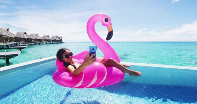 Phone - Travel vacation woman on inflatable Flamingo float mattress using app on mobile cell phone in swimming pool, maybe for booking hotel while relaxing sunbathing on resort pool in bikini