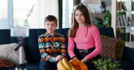 Caucasian family kids brother and sister startting to record online video at home. Two digital modern children siblings recording videovlog in living room.