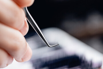Fake black long eyelash extender holds pair of tweezers while working - obrazy, fototapety, plakaty