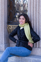 Vertical shot of beautiful smiling middle eastern young woman with long brown hair in casual warm clothes sitting on front step leaning against patrimonial building, Old Quebec, Quebec, Canada