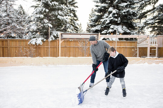 Father And Snow Shoveling Snow Off Backyard Ice Hockey Rink
