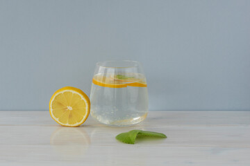 Transparent glass of water, round lemon slice stand near and mint leaf lie on light grey and wooden...
