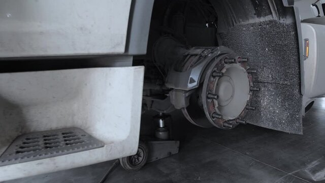 A white truck without front wheel stands on a hydraulic jack at the workshop. Zoom in on the truck's front wheel hub. The mechanic passes in front of the camera