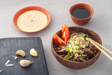 Soba with mushrooms and sesame seeds in a plate of coconut shells on a gray concrete background near garlic and soy sauce and sticks.