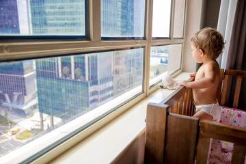 a charming toddler looks out the window at the metropolis while standing in a crib. baby in a crib looks out the window at the city