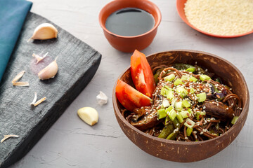 Soba with mushrooms and sesame seeds in a plate of coconut skrlup on a gray concrete background next to garlic and soy sauce.