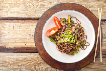 Soba with mushrooms and sesame seeds in a plate on a wooden stand on the table next to chopsticks.
