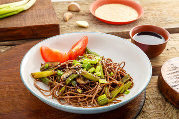 Soba with mushrooms, green beans and sesame seeds in a plate on a stand on a wooden table near chopsticks and ingredients.