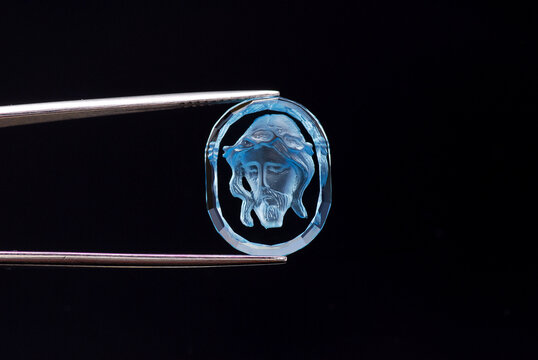 Natural swiss blue topaz oval shape gemstone intaglio cut form of jesus zrist holded in tweezers on black background. Clean transparent gemstone, treated using classic  irradiation method.