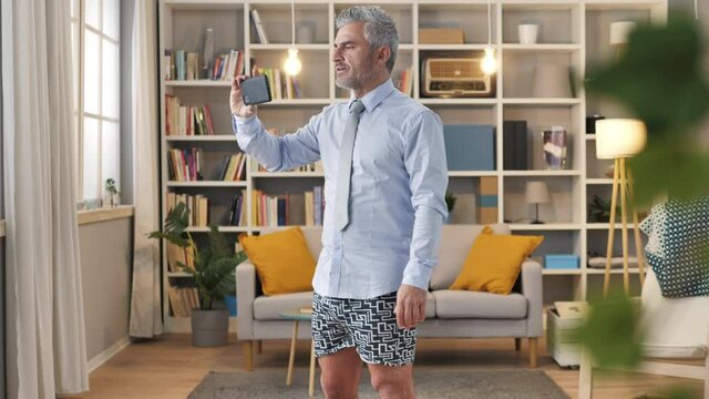 Businessman in shirt and underwear standing in living room makes a business video call,man wearing boxer shorts having a work conversation at the phone from home,funny remote working