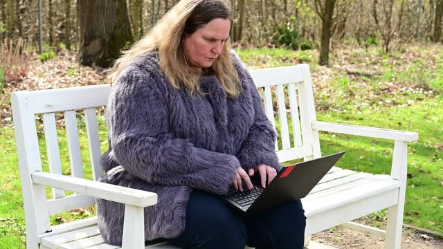 Frau in einer Kaninchenfell Jacke arbeitet im Garten mit einem Notebook im Homeoffice  