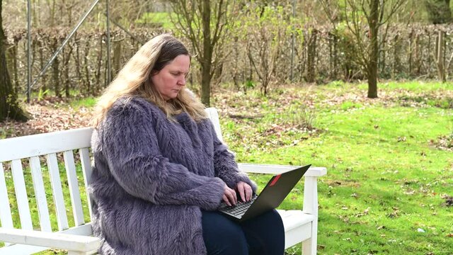 Frau in einer Kaninchenfell Jacke arbeitet im Garten mit einem Notebook im Homeoffice  