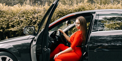 A beautiful young girl in a red overalls sits behind the wheel of a black car on an empty road in the forest