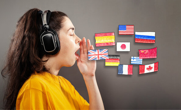 Online language learning. Portrait of a young woman wearing headphones, her hand over her open mouth. Gray background with flags of different countries. The concept of learning foreign languages