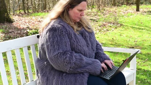 Frau in einer Kaninchenfell Jacke arbeitet im Garten mit einem Notebook im Homeoffice  