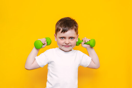 A Little Cute Boy In A White T-shirt With Green Dumbbells Smiles  Isolated On Bright Color Yellow Background. A Sporty Caucasian Kid Does Exercises With Dumbbells. Children's Fitness. Wellness