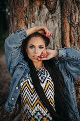 A beautiful girl in a colored dress with bright makeup stands against the background of a coniferous tree trunk. Hair with afro braids.