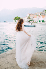 The bride stands on the pier near the old town of Perast and holds the edge of her skirt