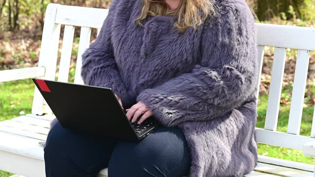 Frau in einer Kaninchenfell Jacke arbeitet im Garten mit einem Notebook im Homeoffice  