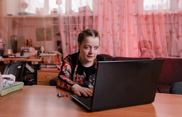 a disabled girl with slanted eyes is sitting at a desk and working on a laptop. training for the...