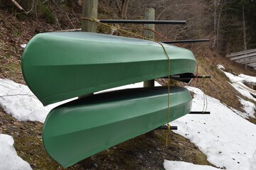  Two green kayaks  stored in racks upside down in side view. They wait for start of water sport season. There are patches of snow around them.