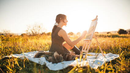Giovane bella ragazza dipinge il quadro su tela seduta su un prato in campagna