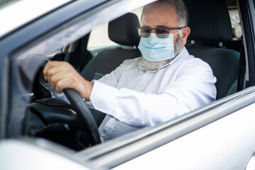 An elderly man in a medical face mask driving a car. Coronavirus pandemic concept. Road trip, travel and old people concept