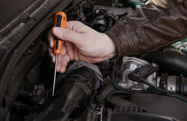A man unscrews the nuts from a spare part in the hood of a car with a screwdriver