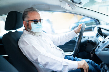 A senior man in a medical surgical mask driving a car. Coronavirus pandemic concept. Road trip, travel and old people concept