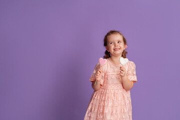 smiling little girl in a soft pink dress holds gingerbread on a stick, in the shape of hearts on a purple background in the studio. The concept of treats for children's birthday and Valentine's Day