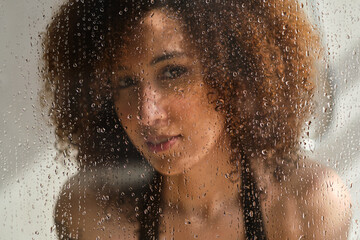 Sensual portrait of young woman taking a shower. Defocused female looks through the glass of the shower stall. Art portrait. Feminine beauty. Skincare wellness.