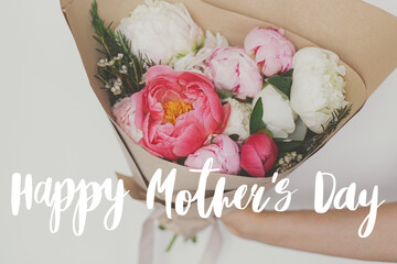 Happy mother's day. Happy mother's day text and hand holding beautiful peonies bouquet in paper on background of white wall. Stylish floral greeting card. Handwritten lettering. Mothers day