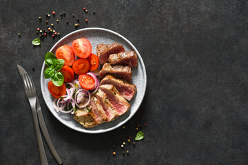 Grilled beef slices and tomatoes and onions in a concrete plate on a black background.