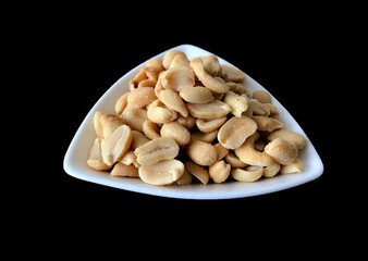 Close up of a heap of salted peanuts on a white triangular dish against a plain black background. No people.