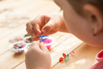 A little girl is engaged in needlework, making jewelry with her own hands, stringing multi-colored...