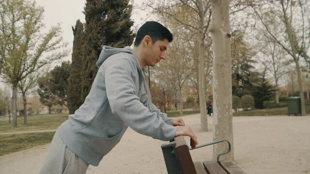 hombre haciendo flexiones apoyado en un banco del parque, hombre de pelo negro con sudadera gris haciendo deporte, hombre haciendo ejercicio en el parque