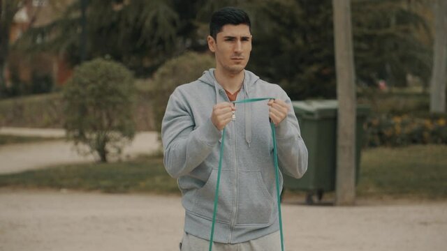 Hombre estirando con banda el&aacute;stica, hombre en sudadera gris calentando antes del entrenamiento, hombre de pelo negro haciendo ejercicio en el parque