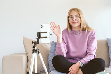 Blogger or vlogger recording video on mobile phone at home and making content for internet. Young attractive woman greeting followers.