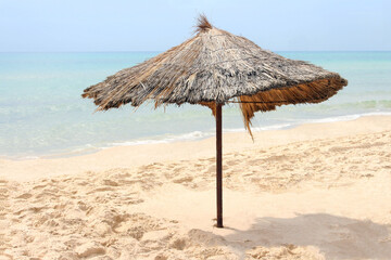 Straw beach umbrella on a tropical sandy beach on a background of blue sea on a sunny day. Idyllic travel and summer vacation concept. Beach umbrella with straw on the sand on a background of blue sea