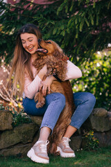 Happy dog licking the face of woman sitting on the yard near tre