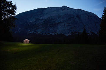Hut in the mountains/Alps/Karwendelgebirge in front of mountain massif. Hut is illuminated orange, mountain is wrapped in a soft blue. Green meadow in the foreground. Night scene. Normal perspective. 