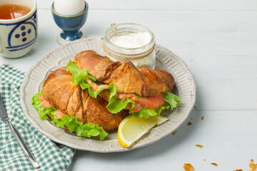 Croissant sandwiches with salmon and green salad, on a gray plate with cream cheese and a slice of lemon. On a gray light background, horizontal with space