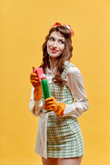 A young female cleaner holds tools for cleaning the house. A bottle of household chemicals and a sponge for washing dishes. He looks at the camera. Pin-up style. Yellow background.