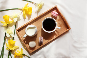 Cup of coffee with milk and French macaroons on a wooden tray and yellow daffodil flowers. Cozy home interior. Spring concept. Breakfast in bed