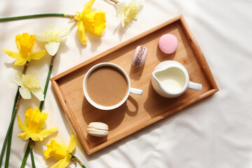 Cozy home interior. Cup of coffee with milk and French macaroons on a wooden tray and yellow daffodil flowers. Breakfast in bed